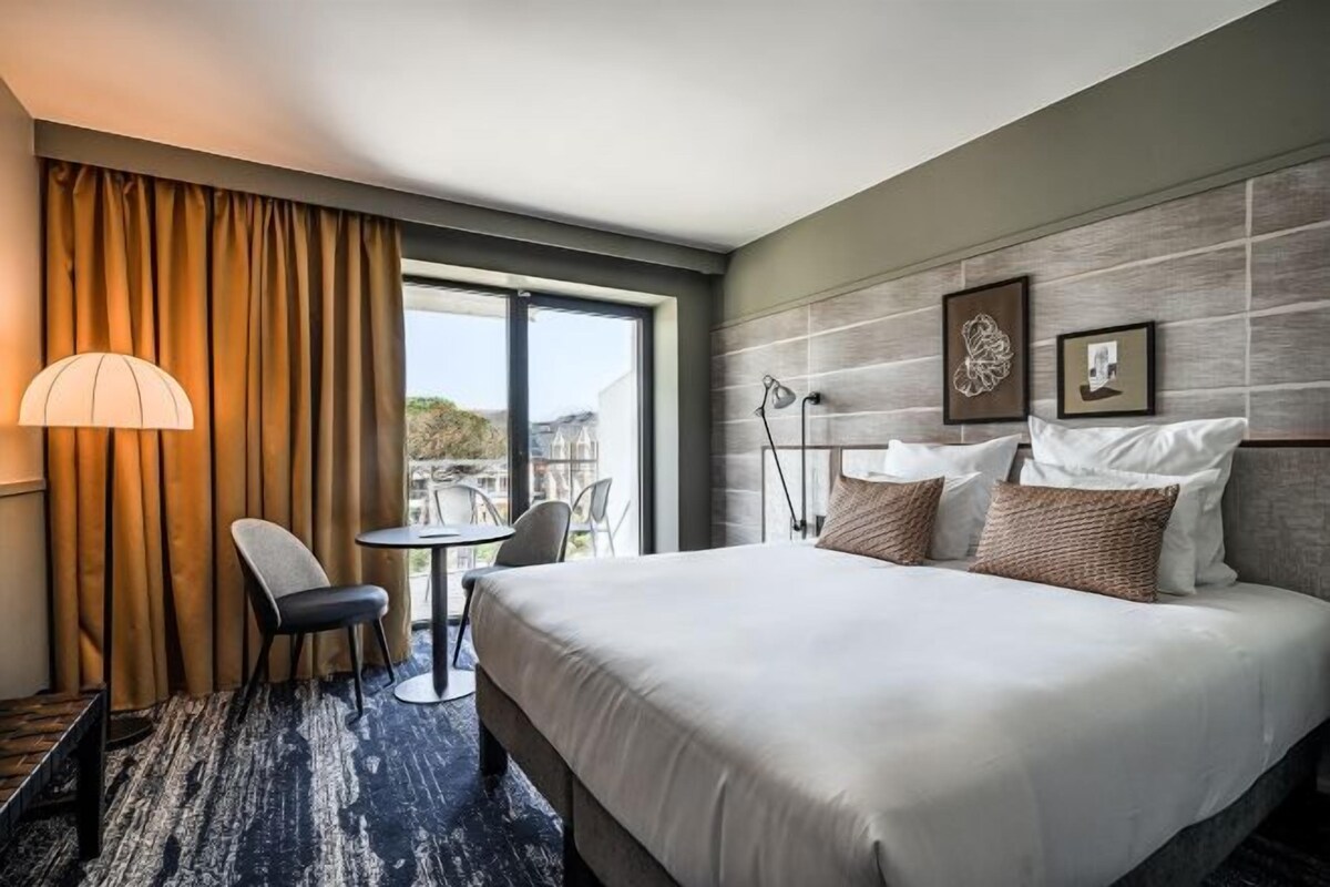 A hotel room with a large bed, a desk, a chair, a lamp, and a view of the outdoors.