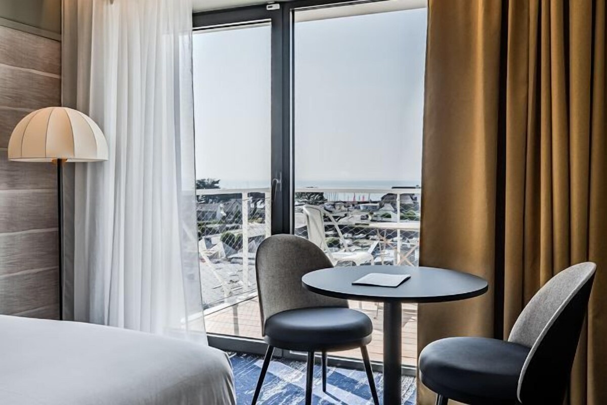 A hotel room with a bed, a table with two chairs, a lamp, and a view of a cityscape through large windows.
