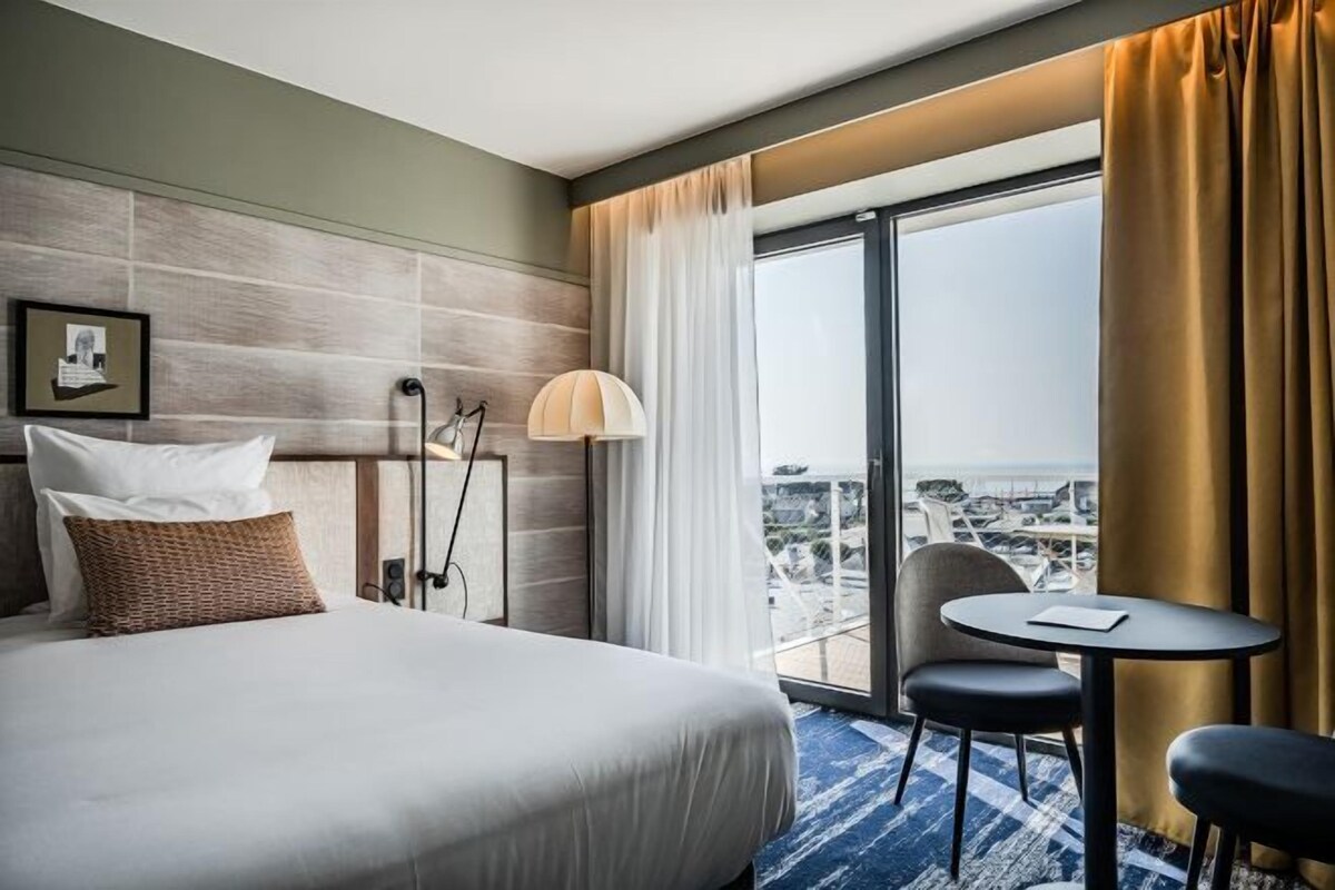 A hotel room with a large bed, a small table, a chair, a lamp, and a view of the city through a large window.