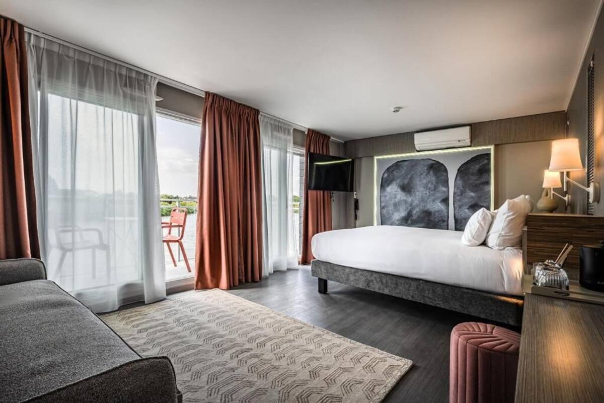 A modern hotel room with a large bed, a sofa, a desk, and a view of the outdoors.