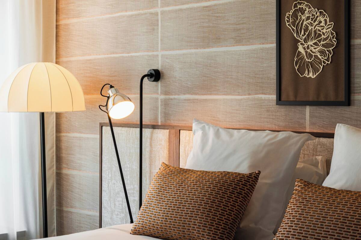 A neatly made bed with patterned pillows, a wall-mounted lamp, and a framed floral artwork.