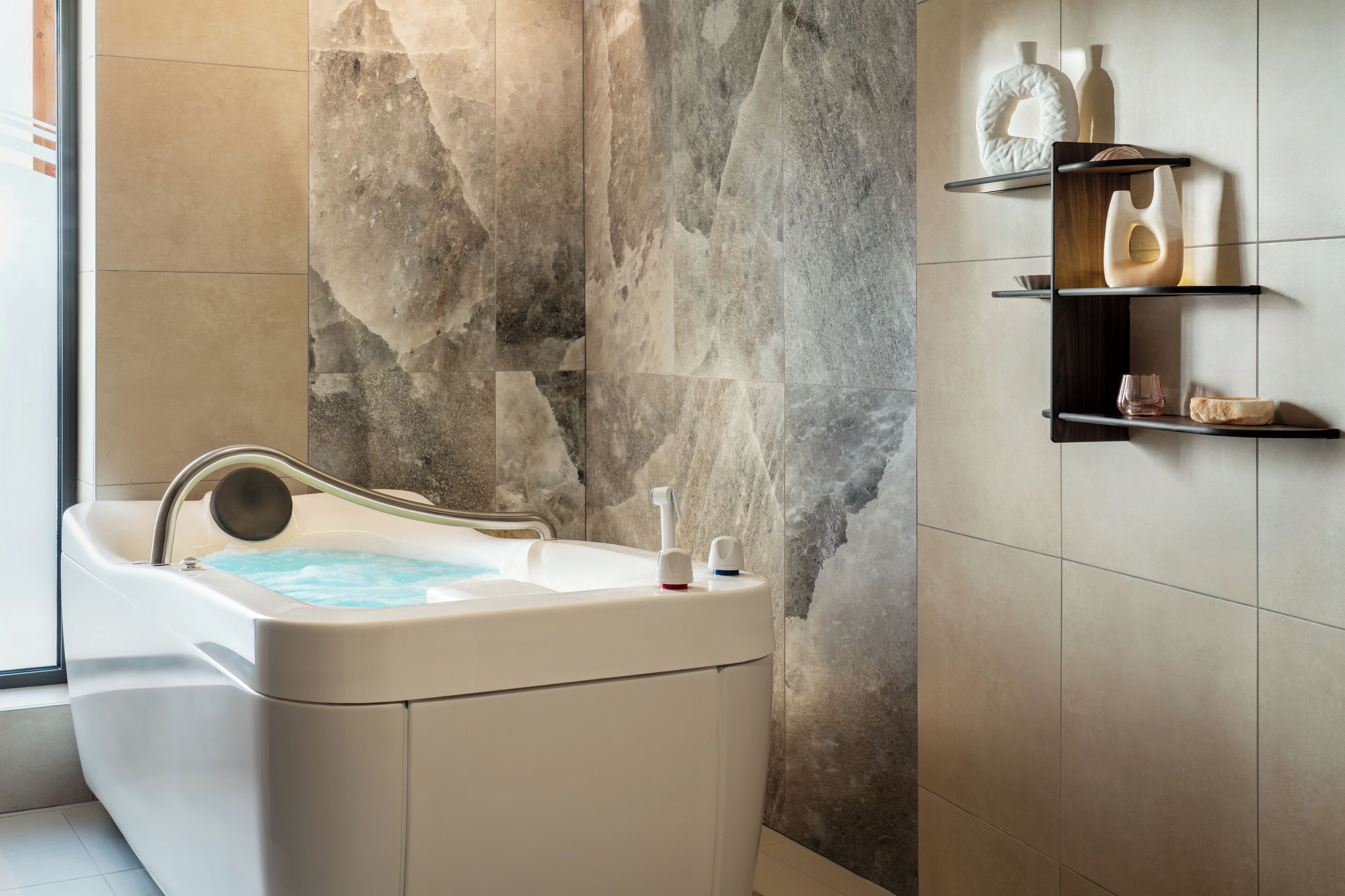 Un baño moderno con pared de piedra, una bañera y estantes minimalistas con objetos decorativos.