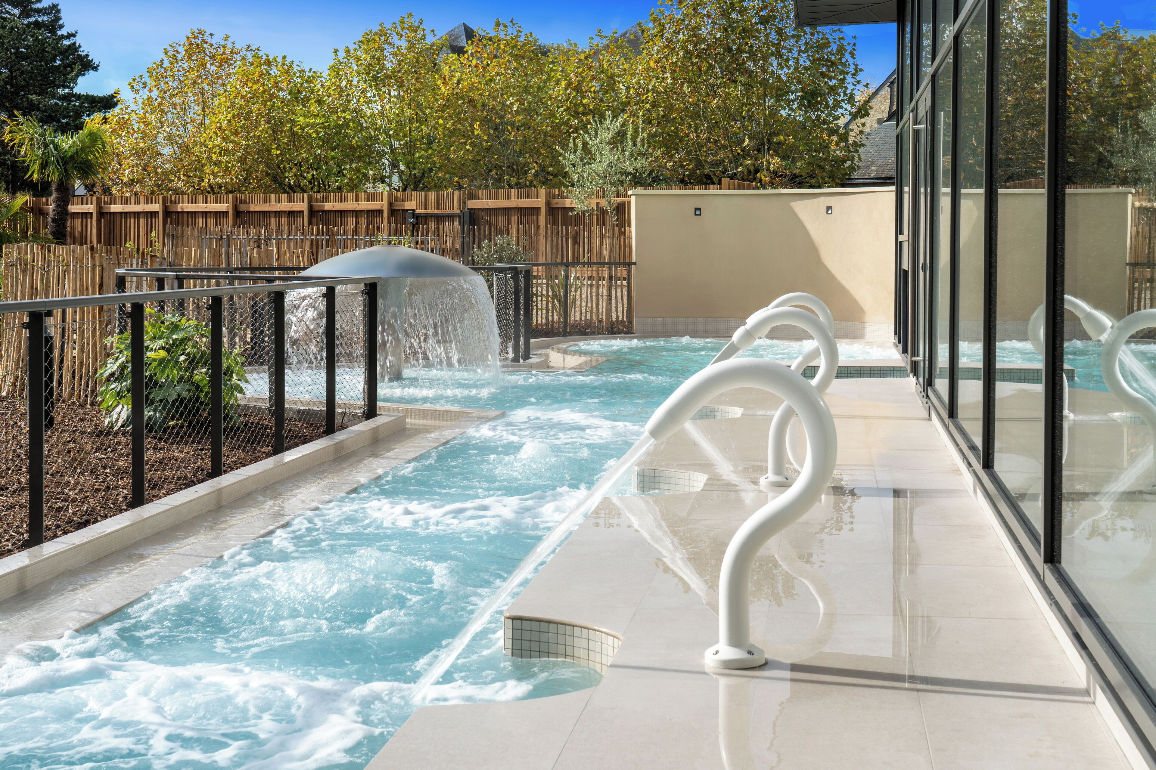 A hot tub area with a wooden fence, a waterfall feature, and a glass railing.