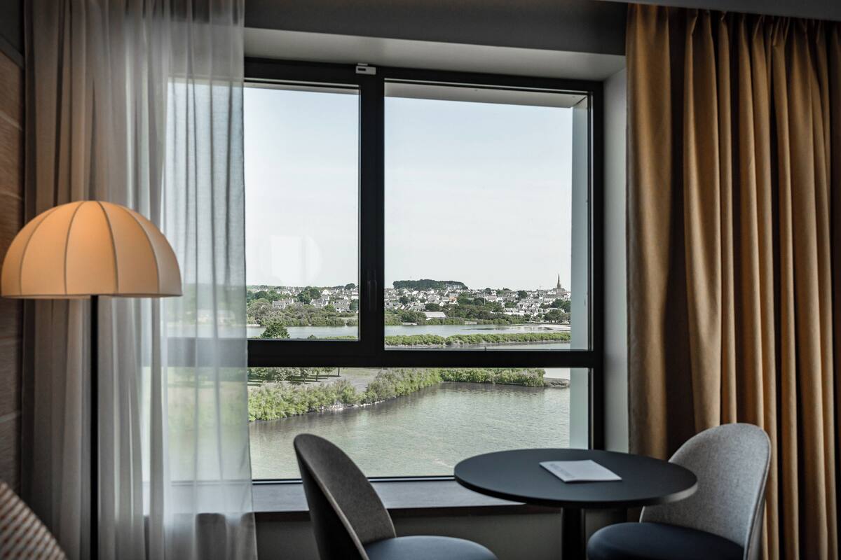 A hotel room with a large window, a view of a river, and two chairs around a small table.