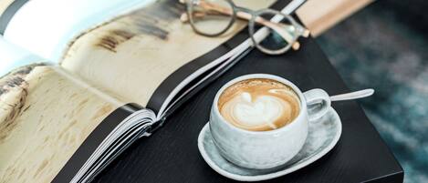 Una taza de café sobre un platillo, junto a un libro abierto con gafas encima.