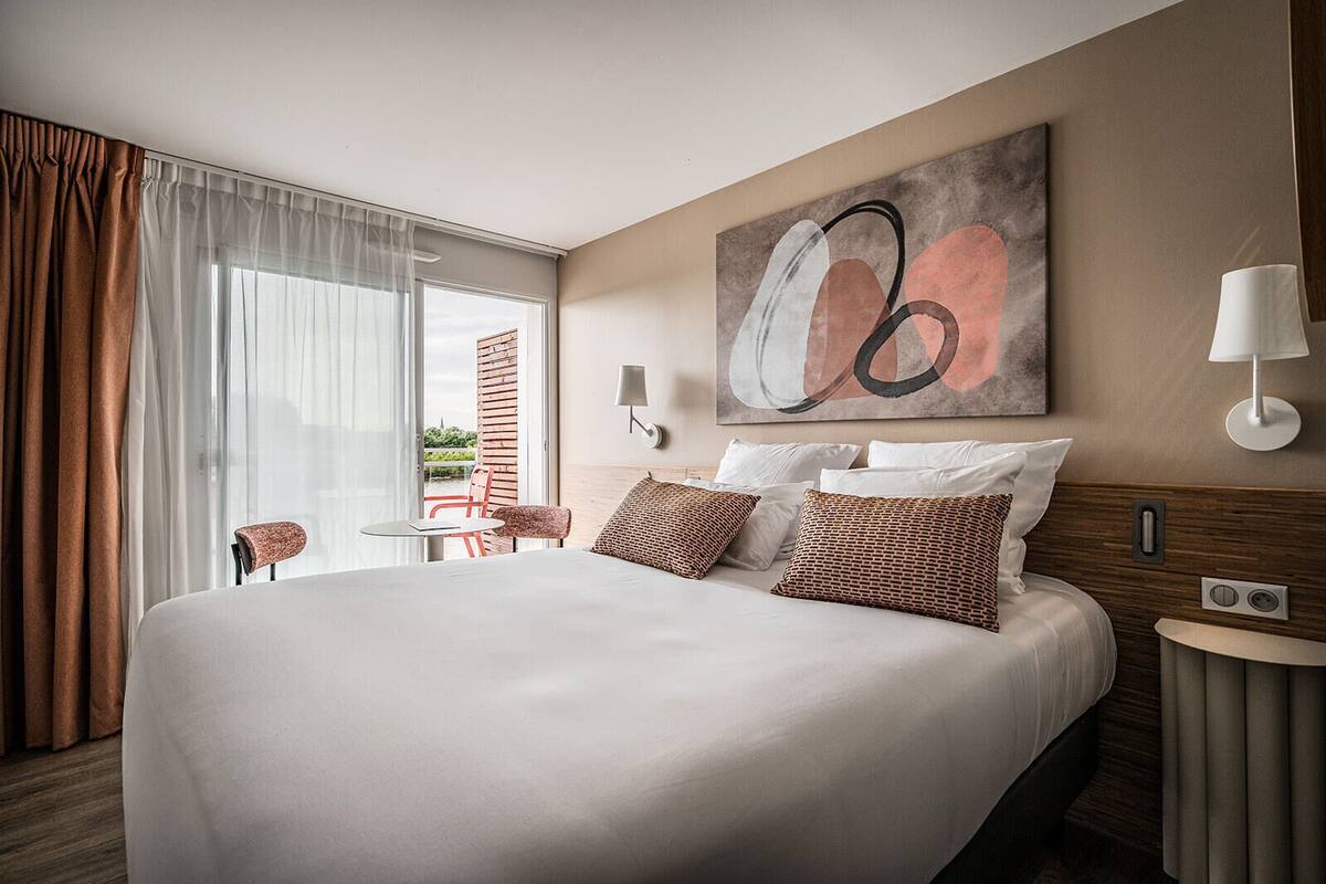 A modern hotel room with a large bed, a small table, a chair, and a view through the window.