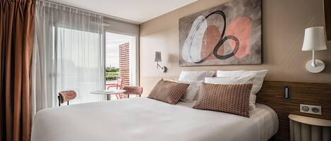 A modern hotel room with a large bed, a small table, a chair, and a view through the window.