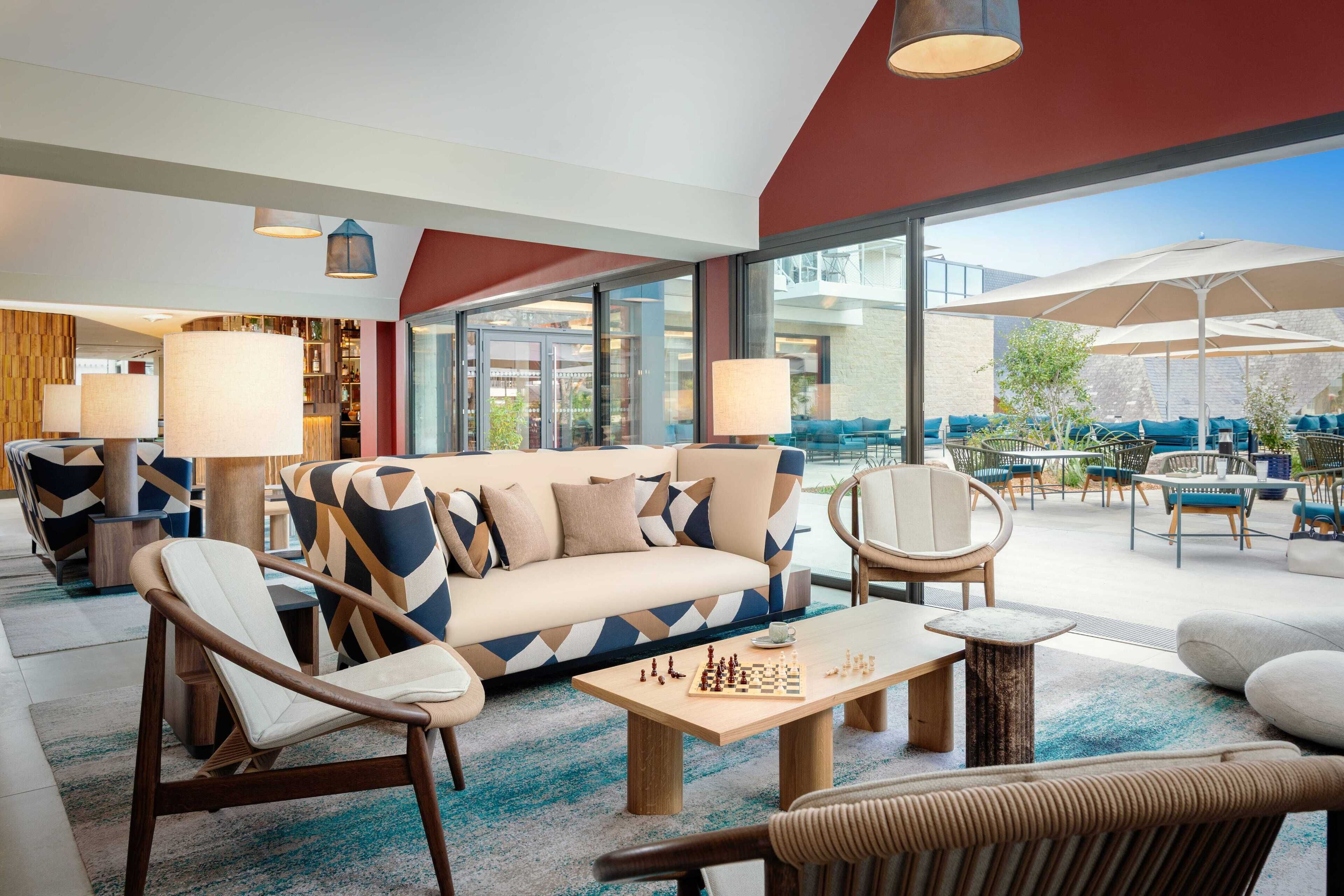 Uma área de lounge moderna com um sofá, cadeiras, mesa de xadrez e mobiliário de exterior com guarda-sóis.