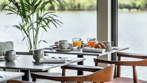 Un desayuno servido en una mesa para dos, con una tetera, tazas y una fuente de croissants. Un amplio ventanal muestra vistas a un lago y follaje.