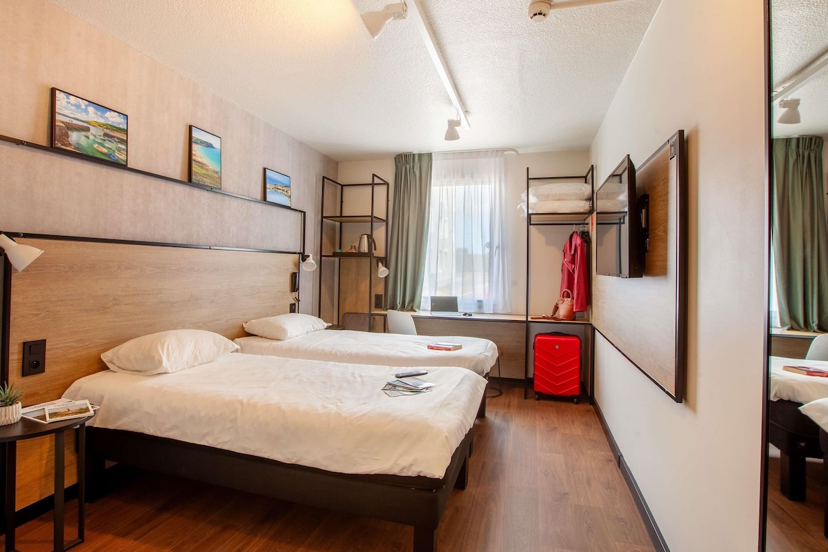 A hotel room with two beds, a desk with a lamp, a small table with a plant, a window with curtains, and a mirror on the wall.