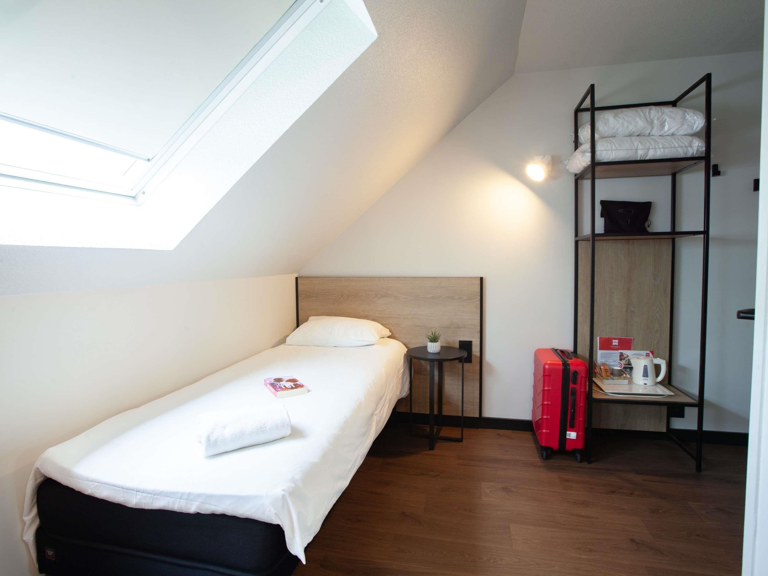 A small, single-bed room with a skylight, a small table with a plant, a red suitcase, and a shelf with a kettle and a coffee pot.