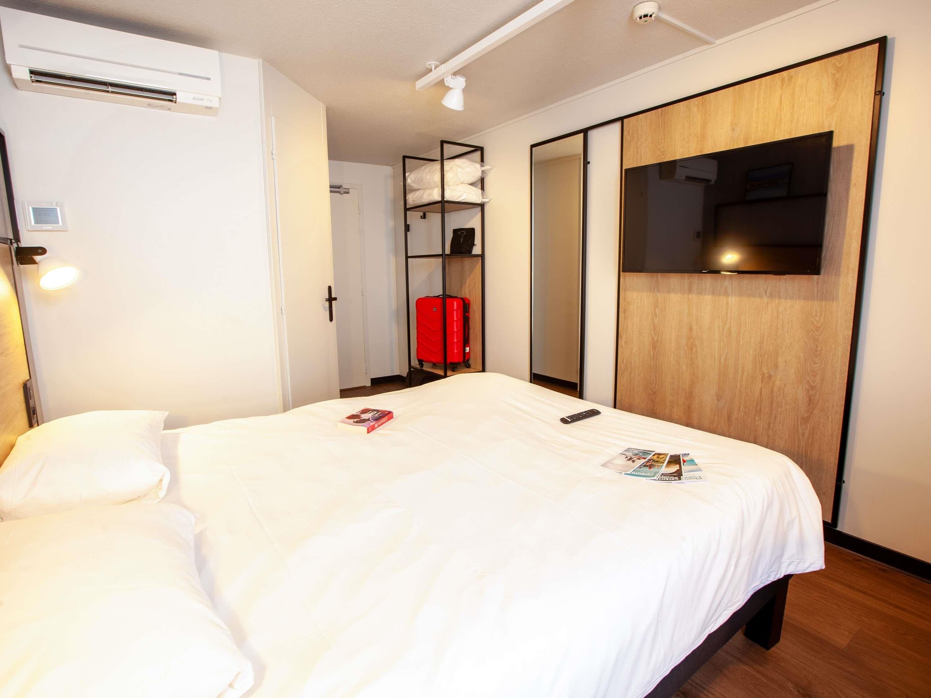 A hotel room with a bed, a wardrobe, a TV, and a red suitcase.
