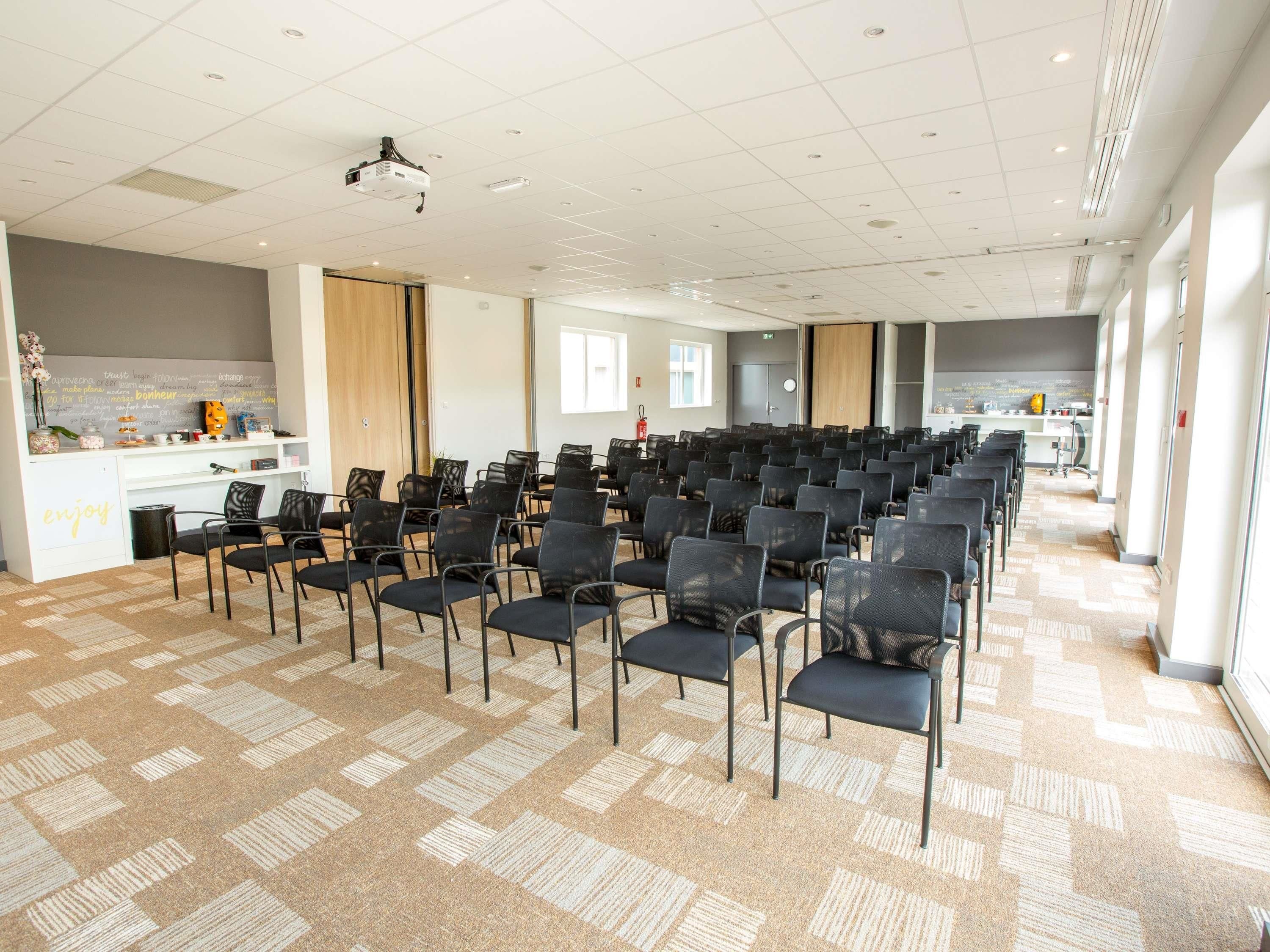 Un salón de conferencias con filas de sillas, un proyector, una pizarra con frases motivacionales y una zona de kitchenette.