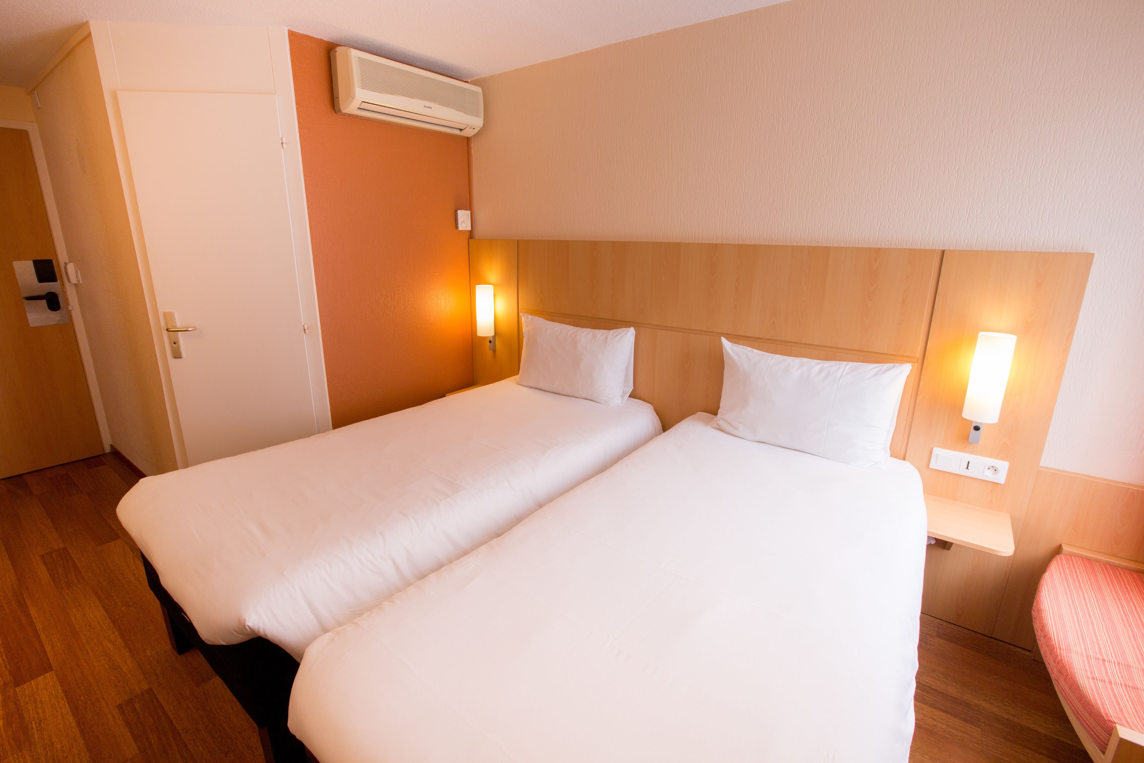 Une chambre d’hôtel avec deux lits simples, une tête de lit en bois, une porte blanche et un climatiseur fixé au mur.
