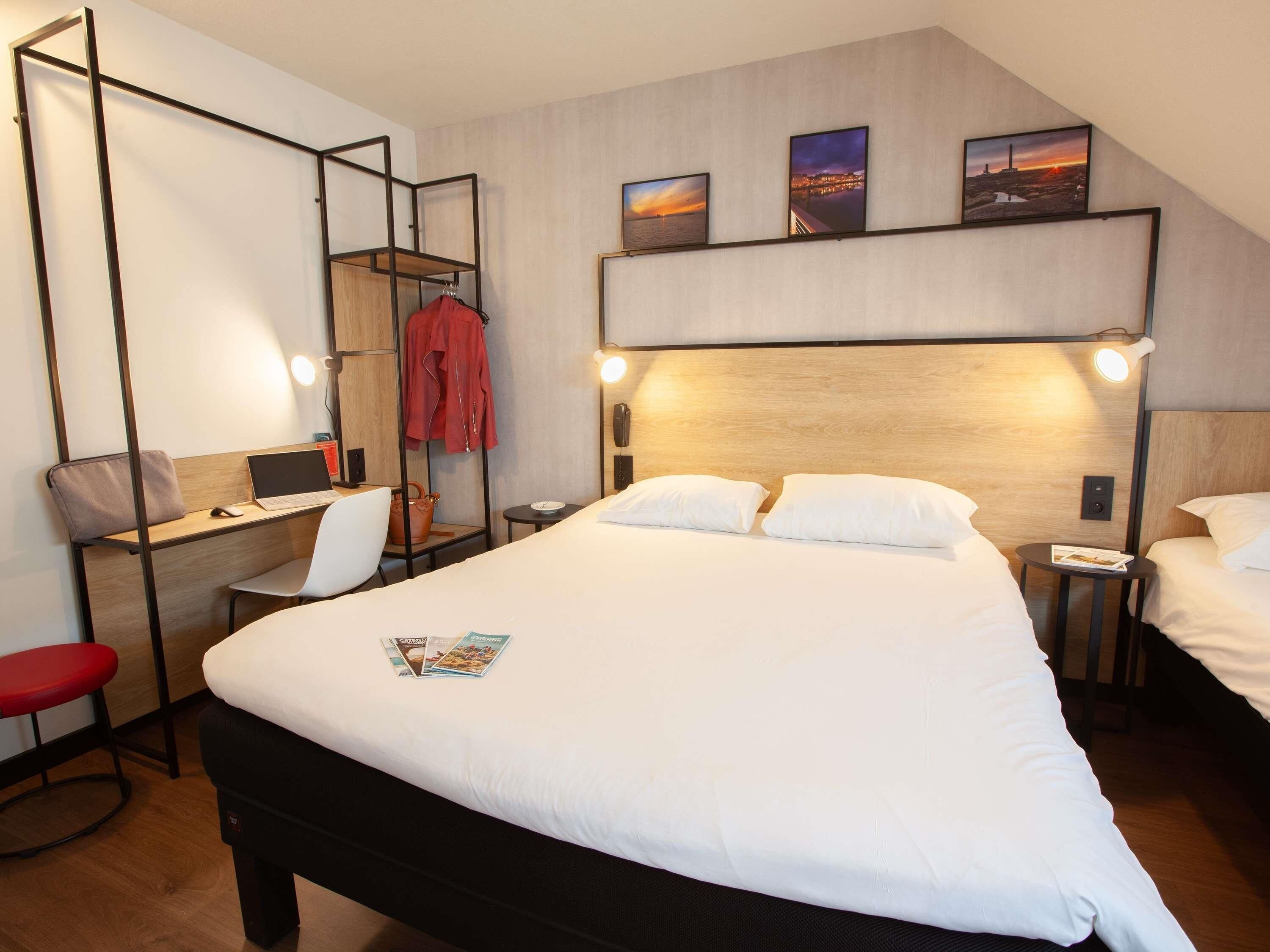 Quarto de hotel moderno com uma cama grande, uma escrivaninha com um laptop, uma cadeira e um casaco vermelho pendurado em um cabide.