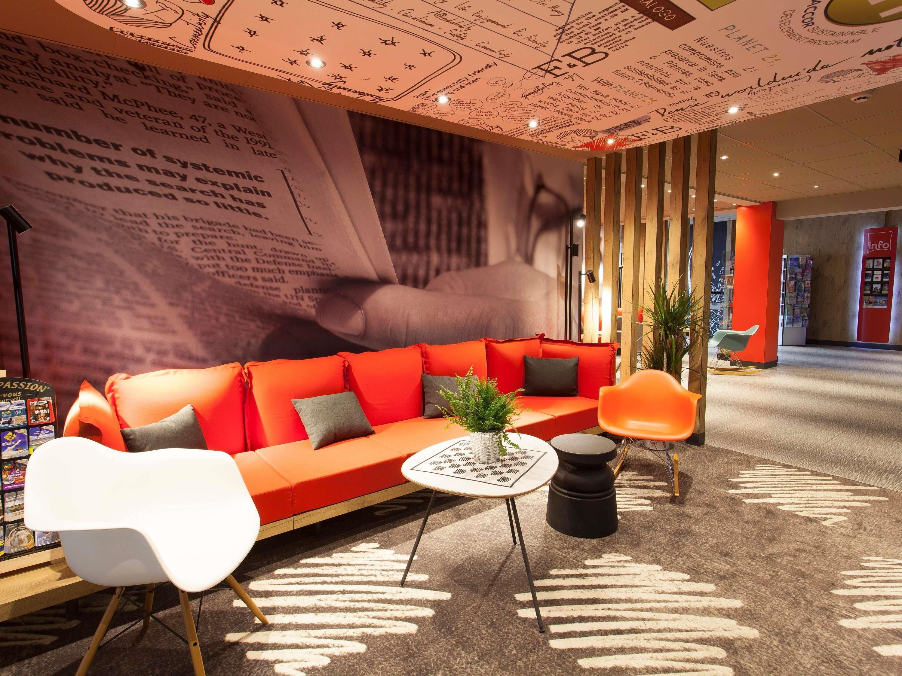 Ein stilvoller Lounge-Bereich mit einem roten Sofa, weißen und orangefarbenen Sesseln, einem kleinen Tisch mit einer Pflanze und einem gemusterten Teppich.