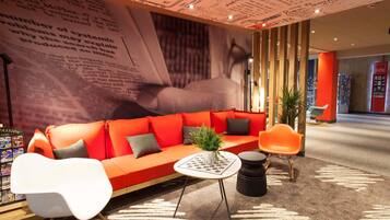 Ein stilvoller Lounge-Bereich mit einem roten Sofa, weißen und orangefarbenen Sesseln, einem kleinen Tisch mit einer Pflanze und einem gemusterten Teppich.