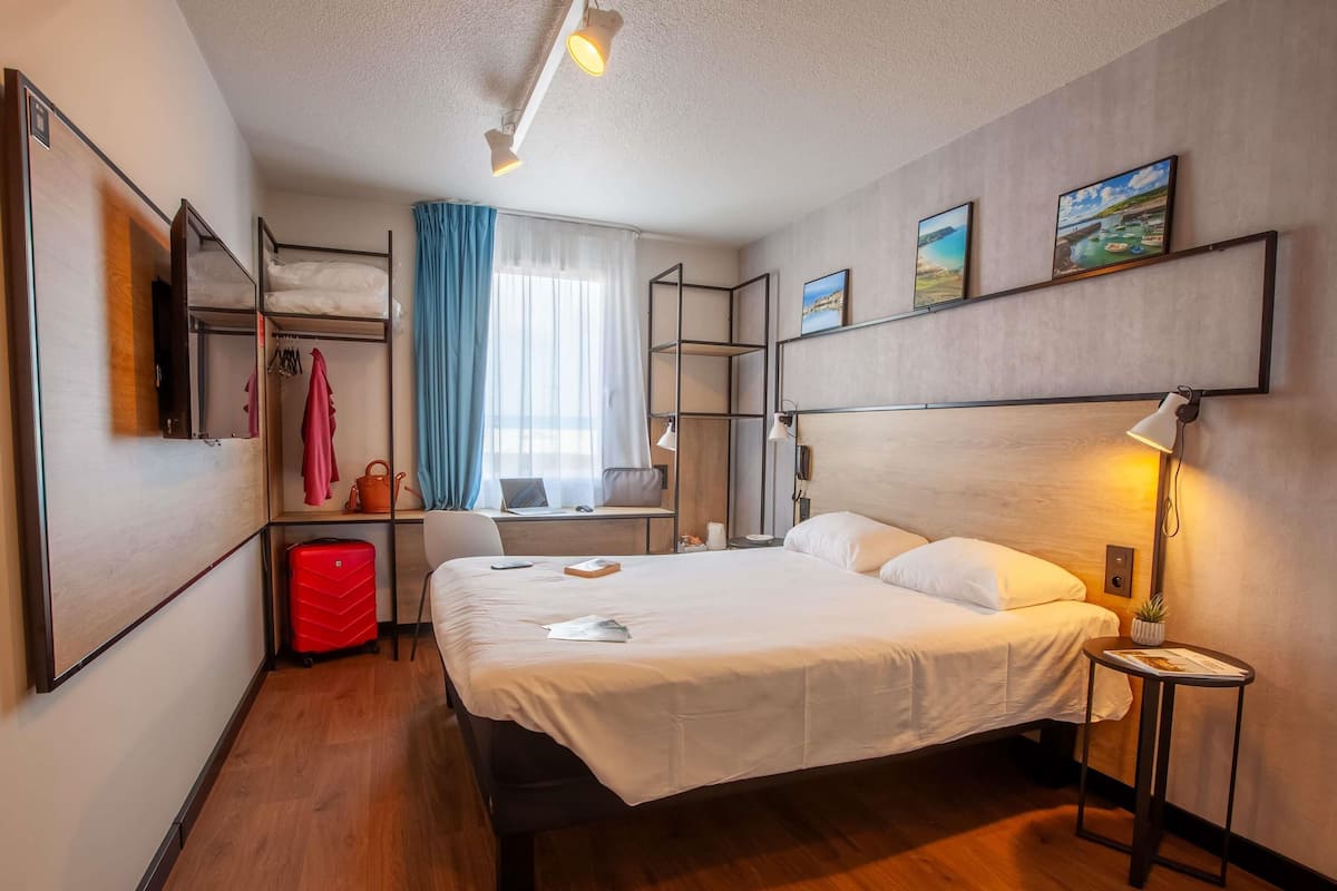 A modern hotel room with a bed, desk, chair, and a small table with a lamp.