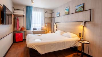 Hotelkamer met bed, nachtkastje, lamp, bureau met stoel, kledingkast en raam met gordijnen.