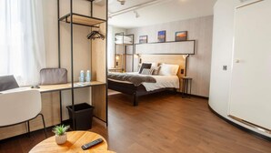 Una habitación de hotel moderna con una cama litera, un escritorio con silla, una mesa redonda de madera y una pequeña planta en maceta.