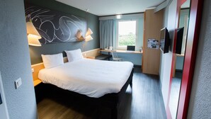A modern hotel room with a large bed, a desk with a computer, a chair, a window with curtains, and a wall-mounted TV. - ibis Cherbourg La Glacerie (Cherbourg-en-Cotentin)