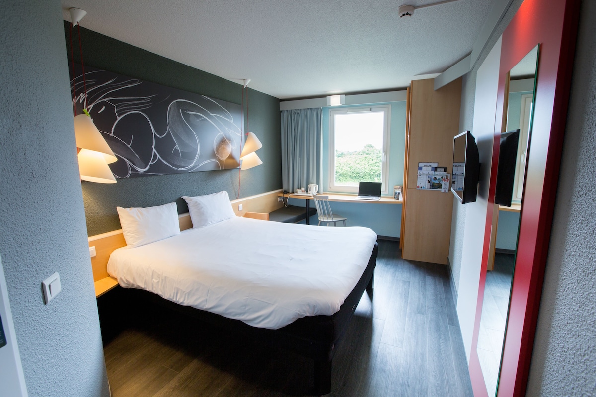 A modern hotel room with a large bed, a desk with a computer, a chair, a window with curtains, and a wall-mounted TV.