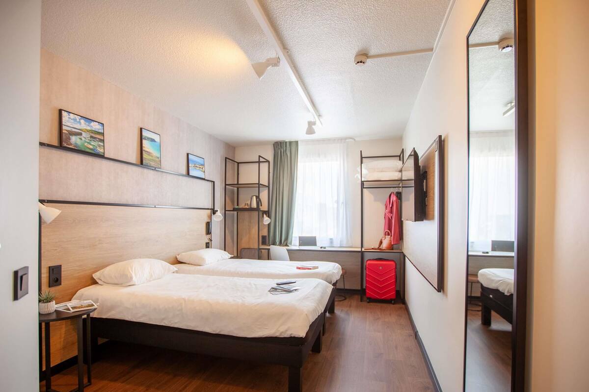 A hotel room with two beds, a desk, a chair, a wardrobe, and a large mirror.