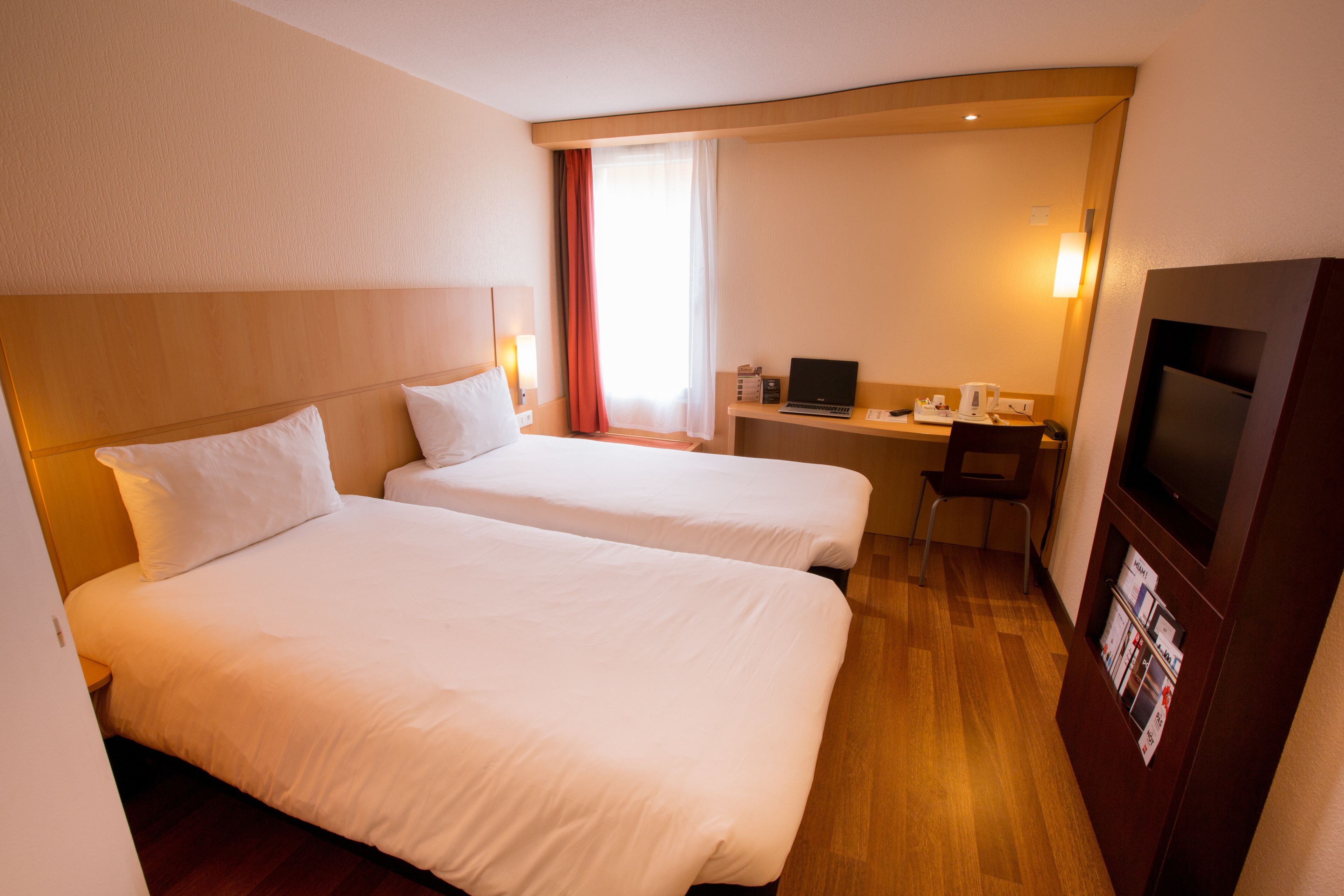 Ein Hotelzimmer mit zwei Betten, einem Schreibtisch mit Laptop, einem Fernseher und einer kleinen Sitzecke.
