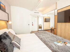 A hotel room with a bed, a TV, a desk, and a door leading to a bathroom. - ibis Cherbourg La Glacerie (Cherbourg-en-Cotentin)