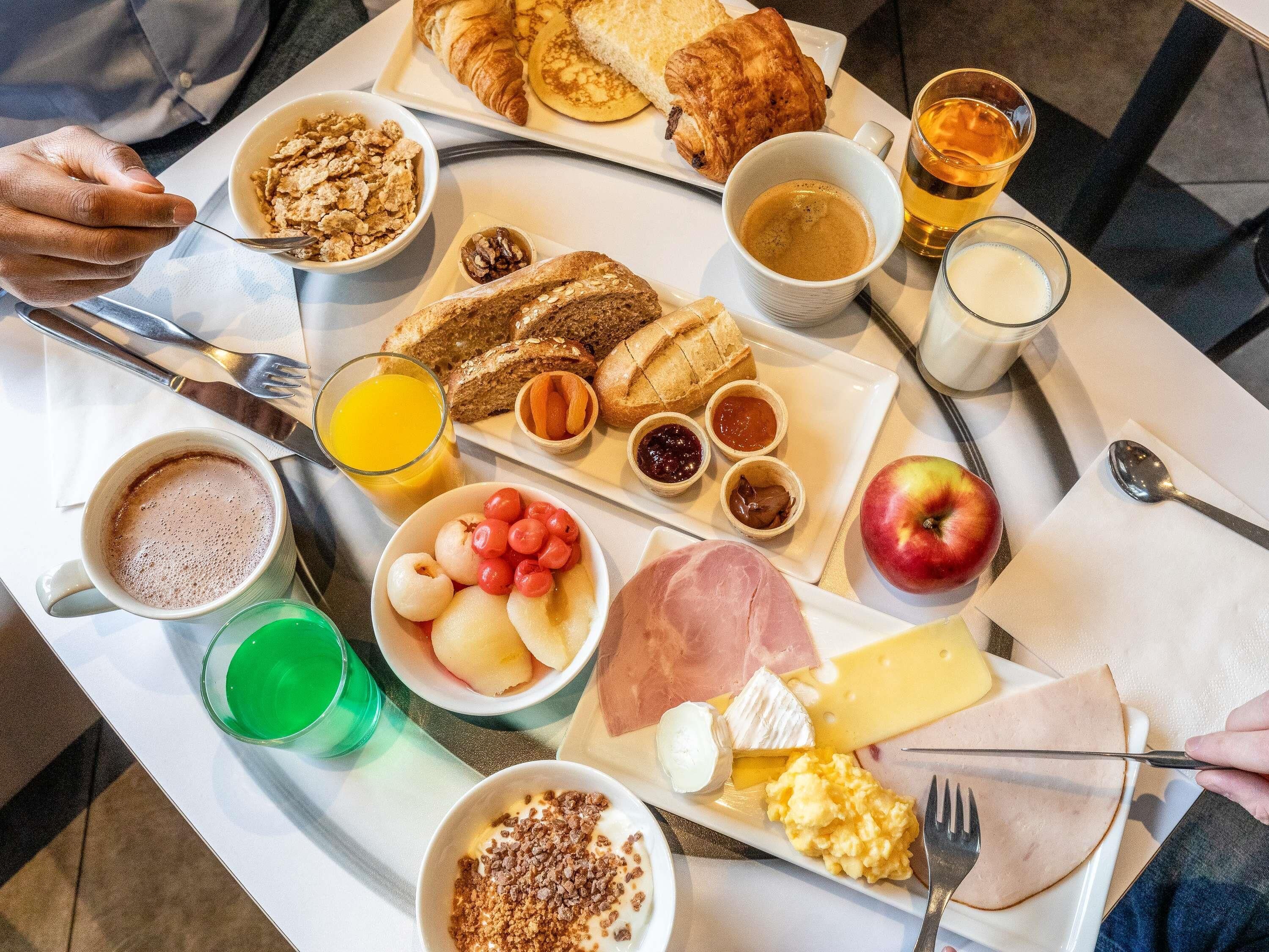 Bandeja de desayuno con cruasanes, pan, queso, jamón, huevos, fruta y bebidas.