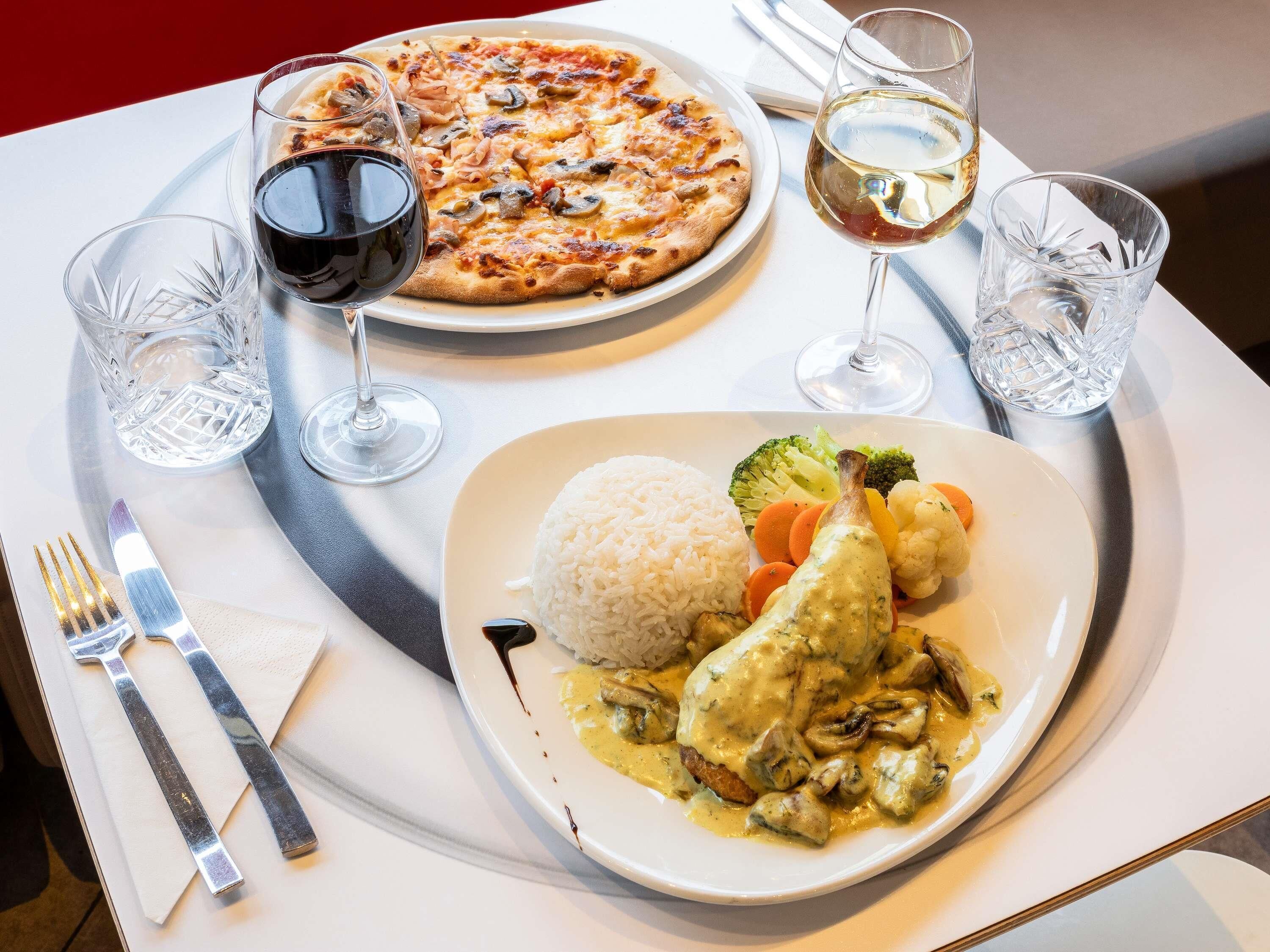 Une table à manger avec une assiette de riz, des légumes et du poulet nappé d’une sauce crémeuse, une pizza et deux verres de vin.