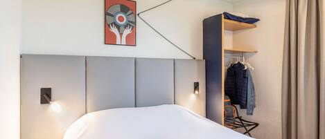 A hotel room with a bed, a headboard, a wall-mounted artwork, a ceiling fan, a small table with a phone and glasses, and a clothing rack.