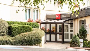 L’entrée d’un hôtel, avec une porte en verre, une enseigne indiquant « Hôtels ibis » et une haie bien entretenue.