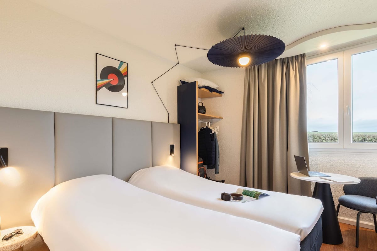 A modern hotel room with a large bed, a desk with a laptop, a chair, and a view of the outdoors.