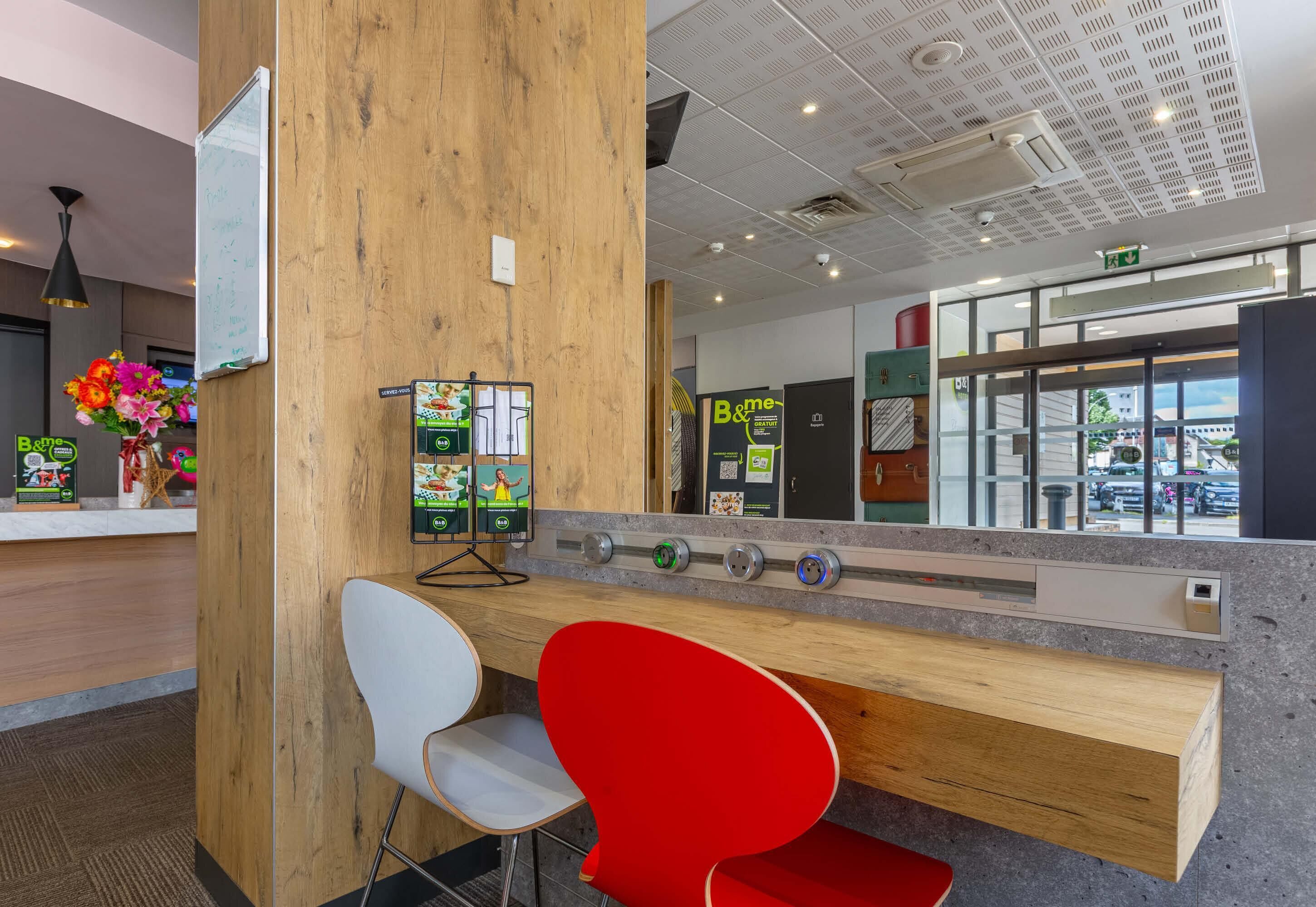 A reception area with a wooden column, a white and red chair, a counter with a digital display, and a view of the outside through glass doors.