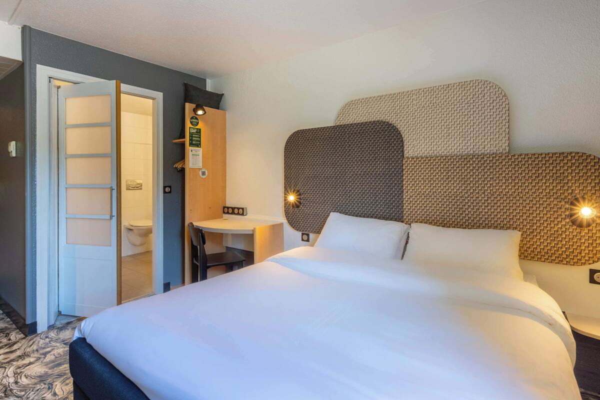 Habitación de hotel con una cama grande, un escritorio pequeño, una silla y un baño visible a través de una puerta abierta.