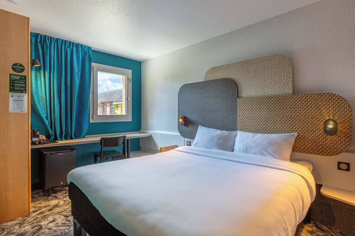 Habitación de hotel con una cama grande, un escritorio y una silla. Se aprecia una ventana con cortinas, una lámpara de pared y un panel de pared texturizado.