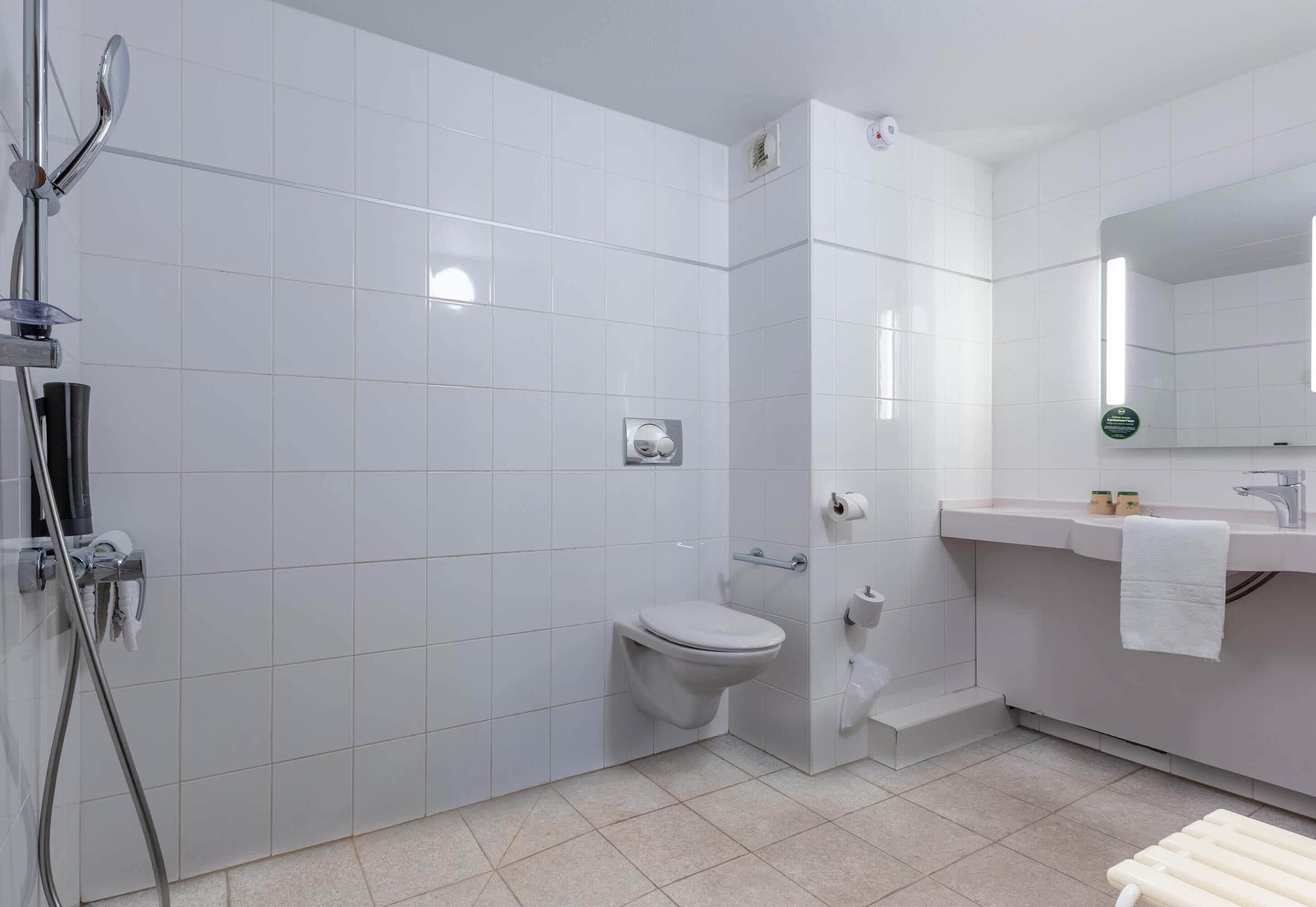 A modern bathroom with white tiles, a toilet, a shower, and a sink with a mirror.