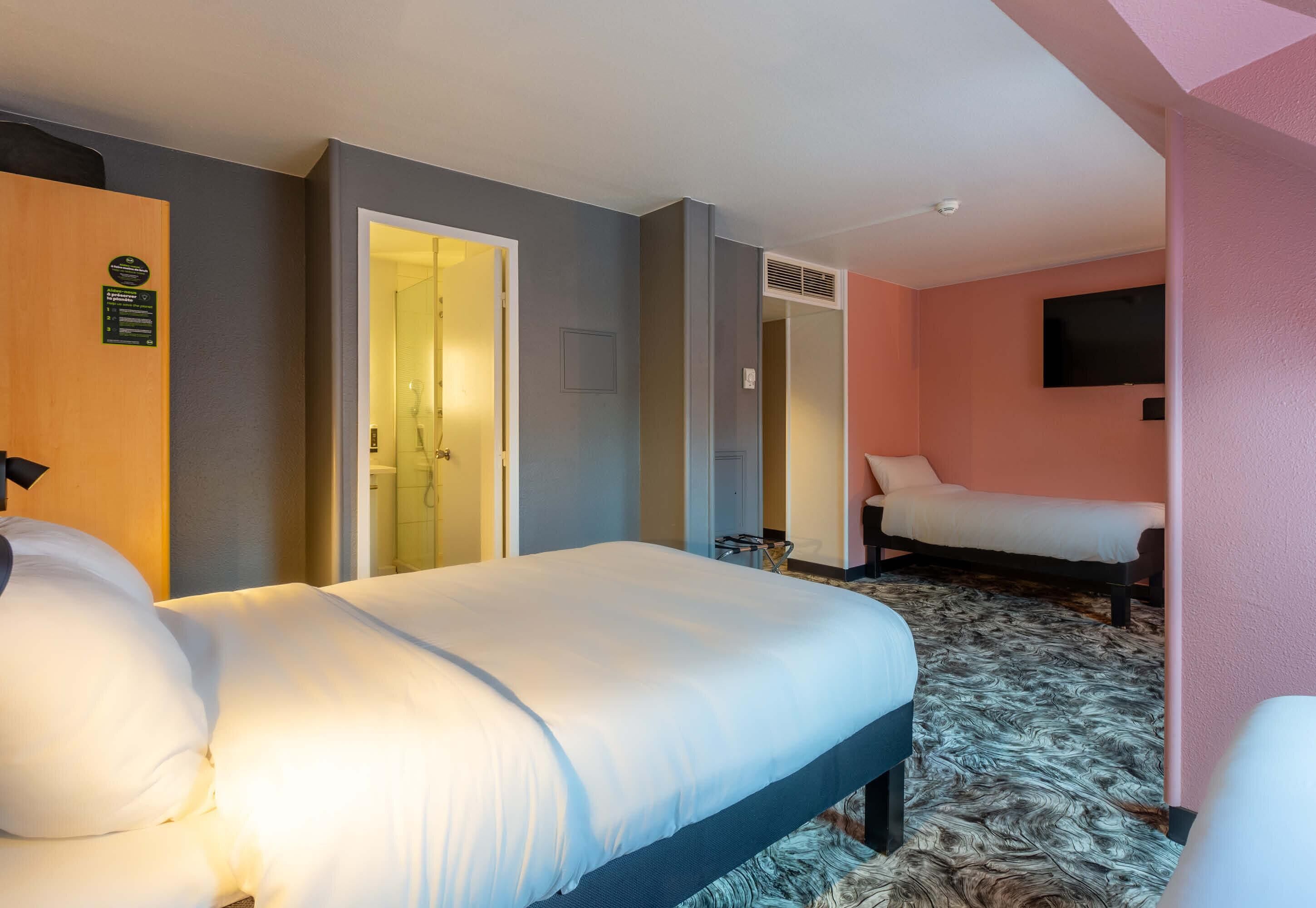 A hotel room with a bed, a second bed, a television, a door, and a shower area.