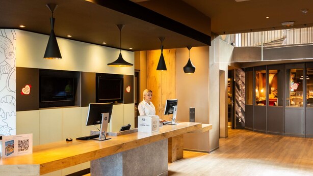 Een receptiebalie van een hotel met een receptionist, een kiosk voor digitaal inchecken en een groot televisiescherm.