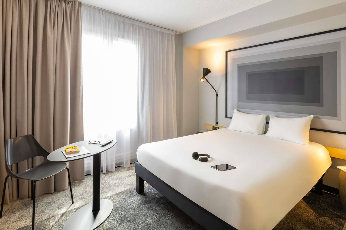 A hotel room with a bed, a desk, a chair, a lamp, and a large window with curtains.