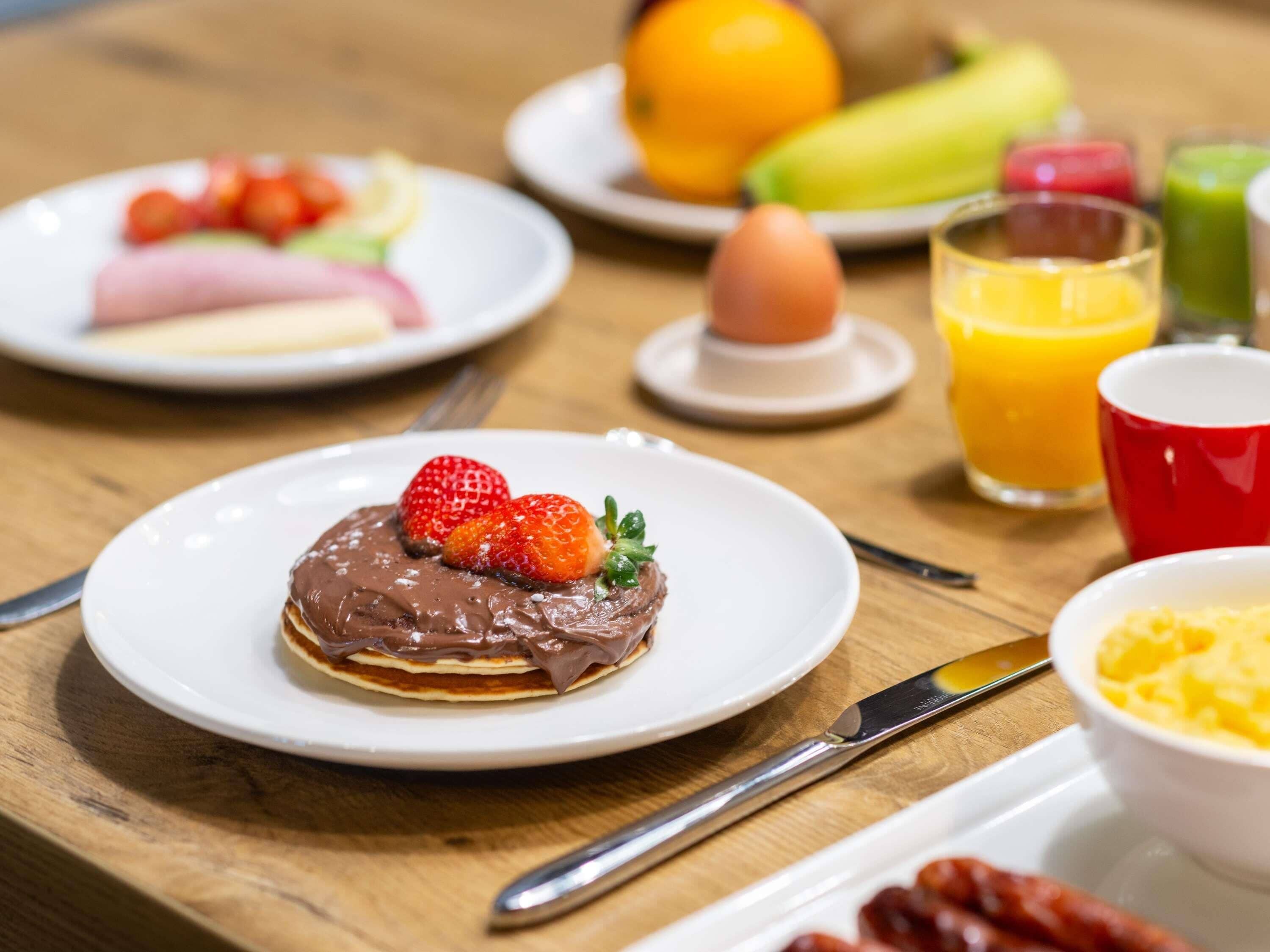 Une table dressée pour le petit-déjeuner avec une assiette de crêpes enrobées de chocolat, garnies de fraises, une assiette de fruits, un verre de jus d’orange et un bol d’œufs brouillés.