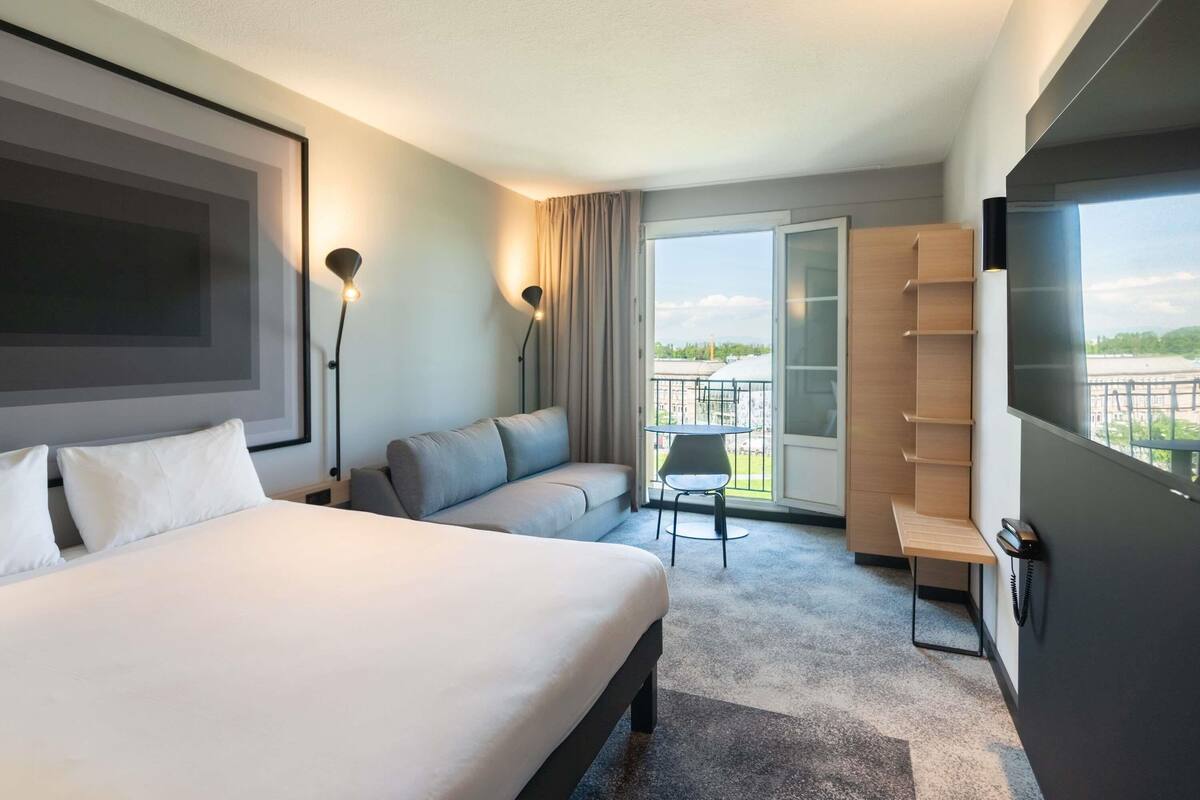 A modern hotel room with a large bed, a sofa, a small table, a chair, and a balcony with a view.