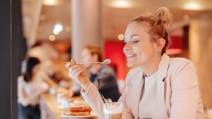 Iemand zit aan een tafel in een café, met een drankje en een lepel.