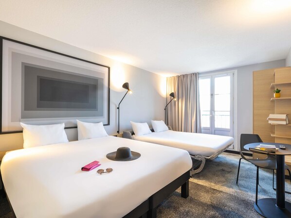 Hotellihuone, jossa on kaksi sänkyä, työpöytä, tuoli, lamppu ja suuri ikkuna, jossa on verhot.