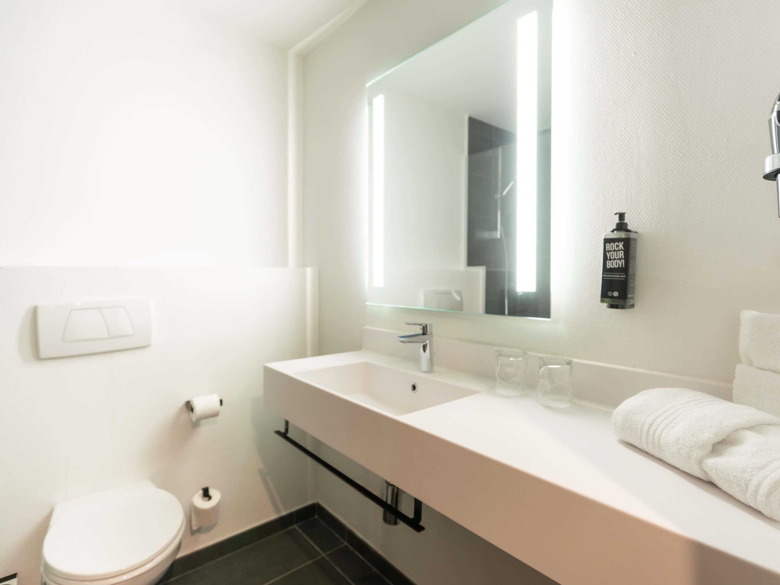 Ein modernes Badezimmer mit einem weißen Waschbecken, einem Spiegel, einem Handtuch und einer Shampooflasche.