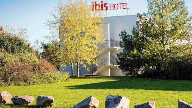 A hotel with a sign reading "ibis HOTEL" in front of a modern building with a balcony. - ibis Mulhouse Ile Napoléon (Sausheim)