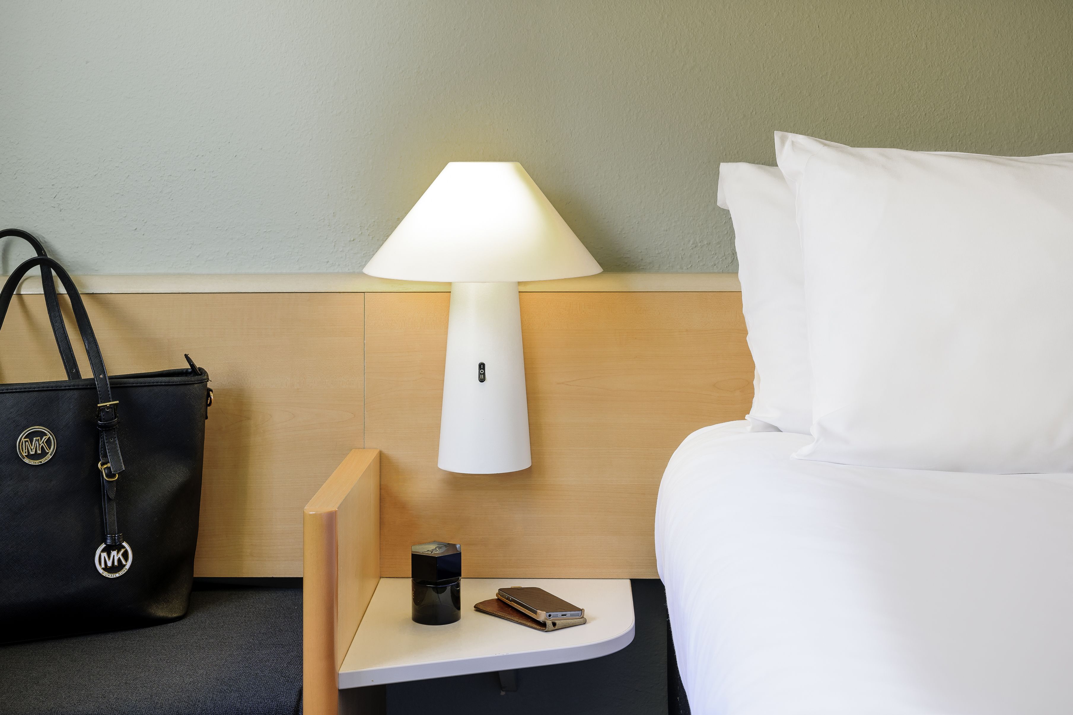 A hotel room with a bed, a nightstand with a lamp, a coffee maker, a smartphone, and a black handbag.