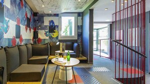 A modern lounge area with a sofa, a table with a drink, and a mural on the wall. - ibis Mulhouse Ile Napoléon (Sausheim)