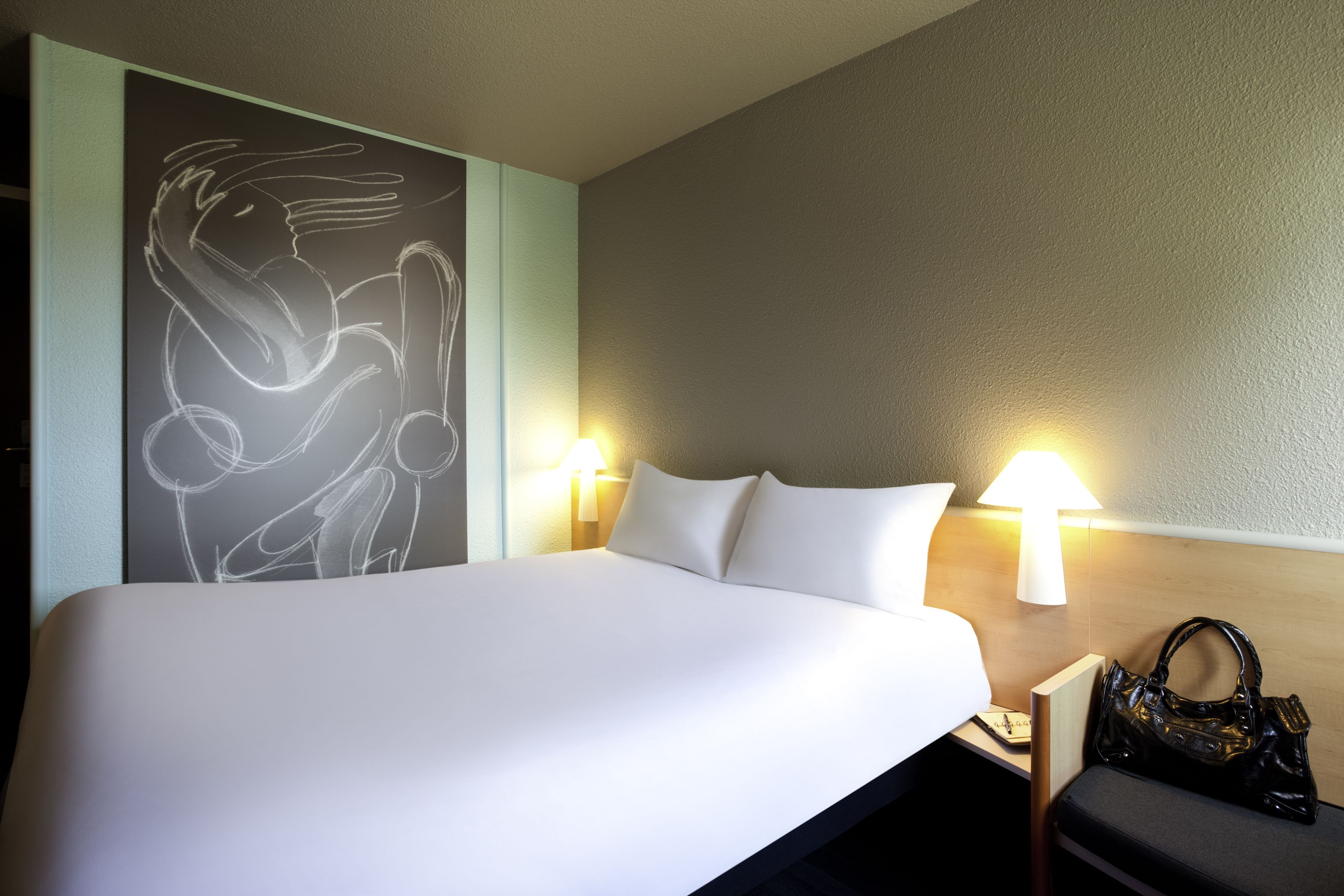 A hotel room with a large bed, two bedside lamps, a wall art piece, and a handbag on the nightstand.